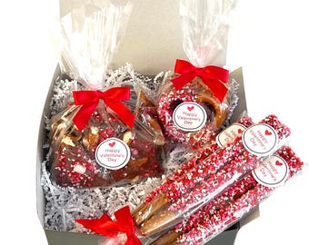 May include: A gift box filled with Valentine's Day treats. The box contains chocolate-covered pretzels, pretzel sticks with red and white sprinkles, and chocolate-covered pretzels with red and white sprinkles. Each treat has a sticker that says "Happy Valentine's Day".