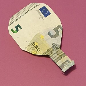 Op de afbeelding: Een briefje van vijf euro gevouwen in de vorm van een peddel. Het briefje is lichtbeige van kleur met een blauw en geel sterrenontwerp. De tekst "ECB ЕЦБ EZB EKP EKT EKB BCE EBC 20" staat bovenaan het briefje gedrukt. De tekst "5" staat in het midden van het briefje gedrukt. De tekst "EURO" staat onderaan het briefje gedrukt.