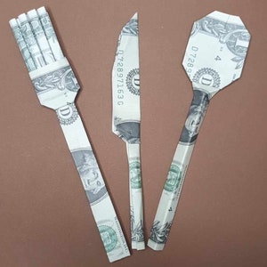 May include: Three origami figures made from US dollar bills, shaped like a fork, knife, and spoon.