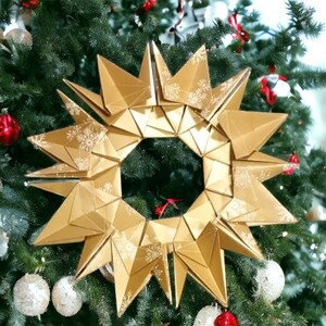 Puede incluir: Un adorno de estrella dorada de origami con un diseño de copo de nieve cuelga de un árbol de Navidad verde.