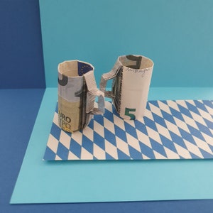 Könnte beinhalten: Zwei Bierkrüge aus gefalteten Fünf-Euro-Scheinen, die auf einer weiß-blauen karierten Oberfläche stehen.