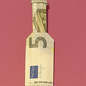 Op de afbeelding: Een briefje van vijf euro gevouwen in de vorm van een fles. Het briefje is lichtgroen van kleur met een afbeelding van een gebouw erop. Op het briefje staat "EKT EKB BCE EBC 2013" en "euro" gedrukt.