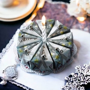 Può includere: Una torta di compleanno fatta di banconote in euro con una candela accesa in cima. La torta è decorata con un pizzo bianco.