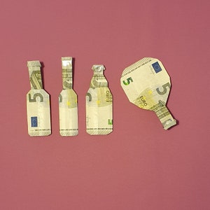 Może przedstawiać: Trzy pięcioeurowe banknoty złożone w kształt butelek i jeden pięcioeurowy banknot złożony w kształt wiosła. Banknoty leżą na różowym tle.