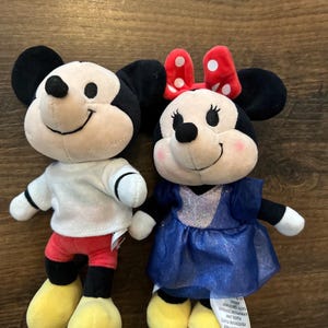 May include: Two plush toys featuring Mickey and Minnie Mouse. Mickey wears a white shirt, red shorts, and yellow shoes. Minnie wears a blue dress with a sparkly bodice and a red bow with white polka dots on her head. Both have black ears.