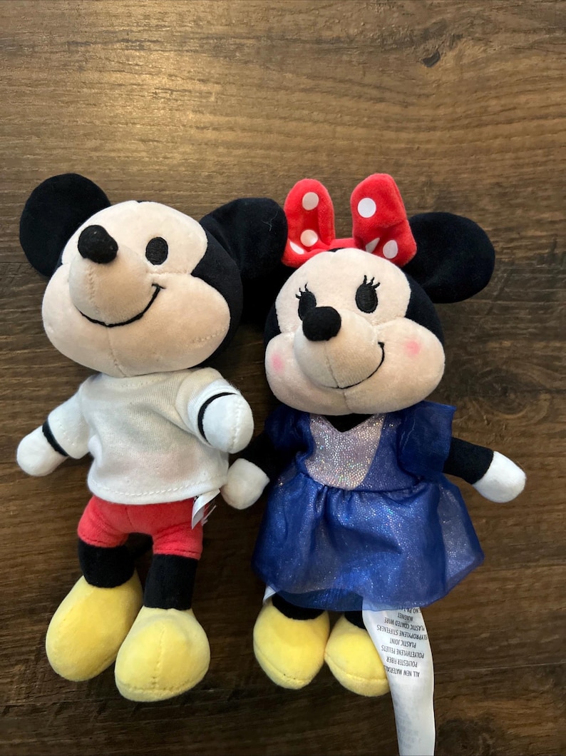 May include: Two plush toys featuring Mickey and Minnie Mouse. Mickey wears a white shirt, red shorts, and yellow shoes. Minnie wears a blue dress with a sparkly bodice and a red bow with white polka dots on her head. Both have black ears.
