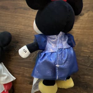 May include: A plush toy of a character with black ears, a red bow, and a blue shimmering dress. The character has white gloves and yellow shoes. A partial view of another plush toy with a white cape and red shorts is visible on the left side.