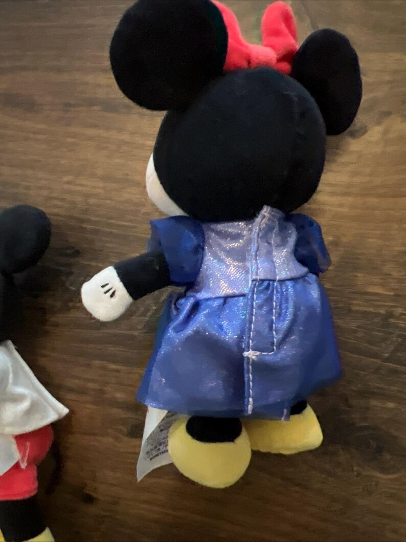 May include: A plush toy of a character with black ears, a red bow, and a blue shimmering dress. The character has white gloves and yellow shoes. A partial view of another plush toy with a white cape and red shorts is visible on the left side.