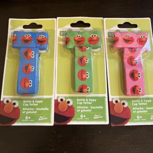 Könnte beinhalten: Drei Flaschen- und Trinkbecherhalter in Blau, Grün und Rosa, jeweils mit einem Elmo-Charakterdesign. Die Verpackung enthält den Text "Bottle & Sippy Cup Tether" und "6+ Monate".