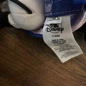 May include: A white plush toy with a black cord and a blue fabric garment featuring a Disney nuiMOs tag. The white care label includes the Disney logo, manufacturing details, and the shopDisney.com website address printed in black text.