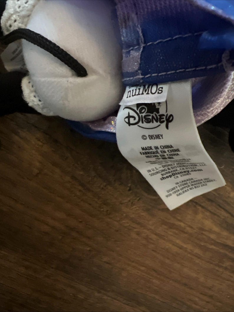 May include: A white plush toy with a black cord and a blue fabric garment featuring a Disney nuiMOs tag. The white care label includes the Disney logo, manufacturing details, and the shopDisney.com website address printed in black text.