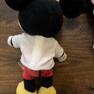 May include: A plush toy featuring a character with large black round ears, a white shirt with a vertical center seam, red shorts, and yellow shoes. The character has black limbs and a small black tail visible at the back of the red shorts.