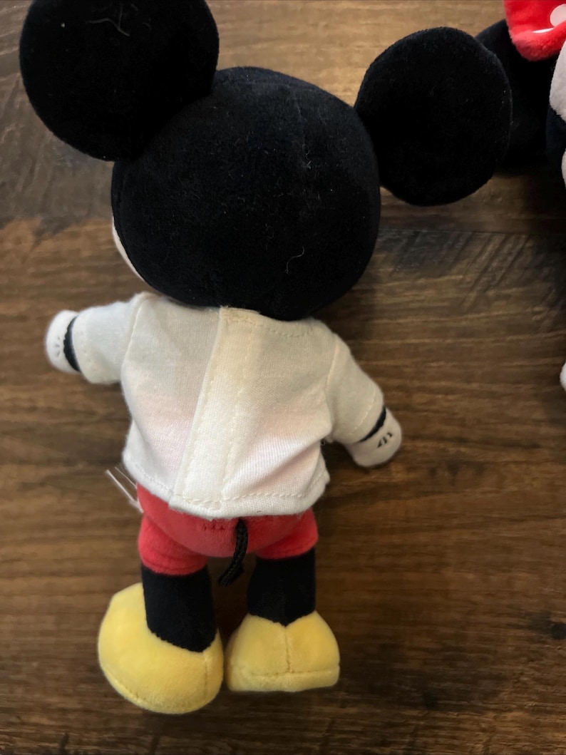 May include: A plush toy featuring a character with large black round ears, a white shirt with a vertical center seam, red shorts, and yellow shoes. The character has black limbs and a small black tail visible at the back of the red shorts.