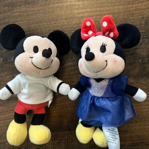 May include: Two plush toys featuring Mickey and Minnie Mouse. Mickey wears a white shirt, red shorts, and yellow shoes. Minnie wears a blue dress with a silver bodice, a red bow with white polka dots, and yellow shoes. Both have black ears.