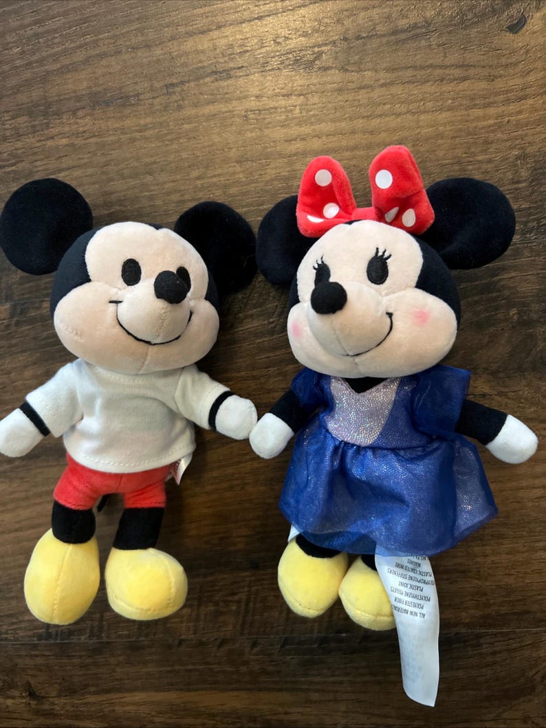 May include: Two plush toys featuring Mickey and Minnie Mouse. Mickey wears a white shirt, red shorts, and yellow shoes. Minnie wears a blue dress with a silver bodice, a red bow with white polka dots, and yellow shoes. Both have black ears.