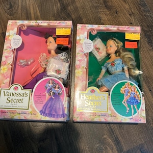 May include: Two dolls from "The Glamour Secrets Collection" are presented in their original packaging. One doll is in a purple dress, the other in a blue dress. The boxes have floral patterns and the text "Vanessa's Secret" and "Heather's Secret".