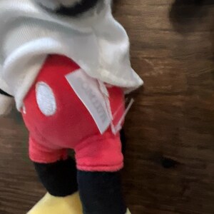 May include: A plush toy character wearing a white shirt, bright red shorts with two white oval buttons, and black legs with large yellow shoes. A white fabric tag is attached to the side of the red shorts, partially obscuring the view of the fabric.