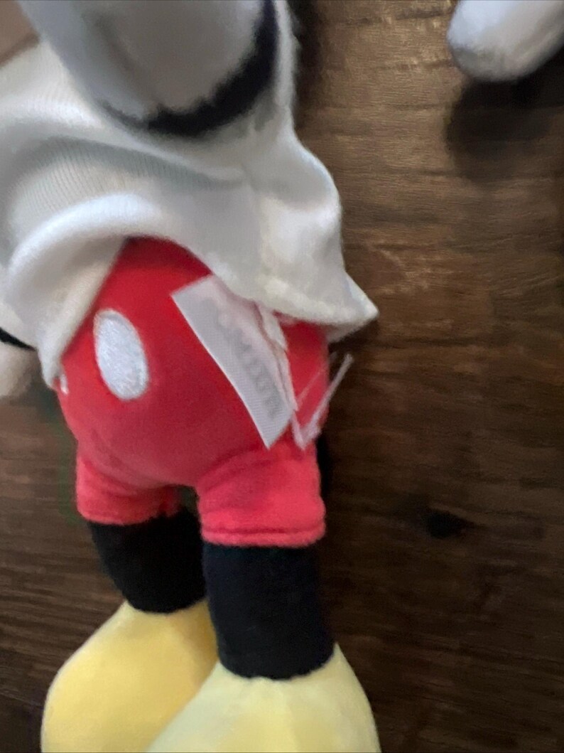 May include: A plush toy character wearing a white shirt, bright red shorts with two white oval buttons, and black legs with large yellow shoes. A white fabric tag is attached to the side of the red shorts, partially obscuring the view of the fabric.
