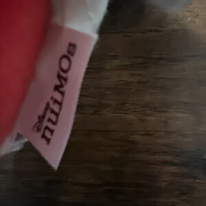 May include: A light pink fabric tag featuring the Disney nuiMOs logo in dark text. The tag is attached to a soft, plush item, with a portion of a red material visible on the left side and a white fabric section visible at the top of the tag.