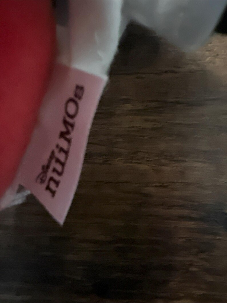 May include: A light pink fabric tag featuring the Disney nuiMOs logo in dark text. The tag is attached to a soft, plush item, with a portion of a red material visible on the left side and a white fabric section visible at the top of the tag.