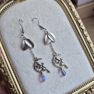 Peut inclure: Boucles d'oreilles pendantes argentées. Elles présentent un motif de feuille en haut, relié à une chaîne avec un croissant de lune et un charm de fée. Chaque boucle a une perle irisée en forme de goutte. Présentées sur une surface crème.