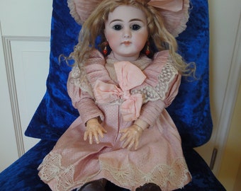 May include: A vintage porcelain doll with blonde hair, wearing a pink dress with lace trim and a large pink hat. The doll has blue eyes and red earrings.