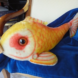 以下が含まれることがあります： オレンジと黄色の大きなぬいぐるみのおもちゃの魚。赤い目と白いお腹をしています。魚は青い毛布の上に横たわっています。