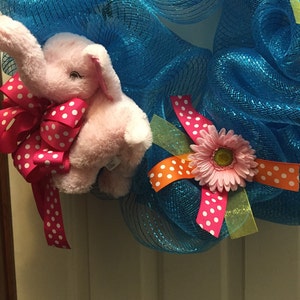 May include: A blue mesh wreath with a pink elephant plush toy, a pink flower with yellow center, and pink and orange ribbon with white polka dots.