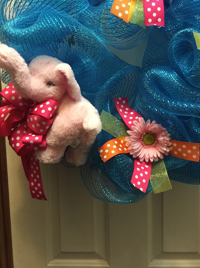 May include: A blue mesh wreath with a pink elephant plush toy, a pink flower with yellow center, and pink and orange ribbon with white polka dots.