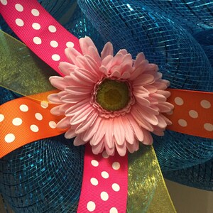 May include: A pink daisy flower with a yellow center is surrounded by three ribbons with white polka dots on a blue mesh background. The ribbons are pink, orange, and yellow.