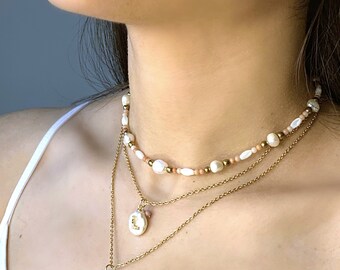 May include: A layered necklace featuring three strands. The top strand has pearls and coral beads. The middle strand has a pendant with the letter "L". The bottom strand has a pink heart pendant. All strands are gold.