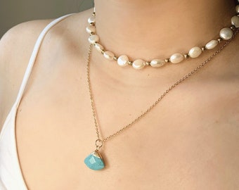 May include: A layered necklace set featuring a pearl choker and a gold chain pendant. The pendant showcases a faceted turquoise stone. The pearls are off-white, and the metal components are gold-toned. The necklace is worn against a white top.