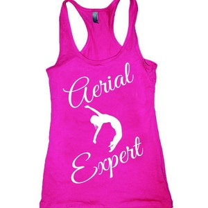May include: A pink tank top with the text "Aerial Expert" in white lettering. A white silhouette of a person performing an aerial maneuver is in the center of the text.
