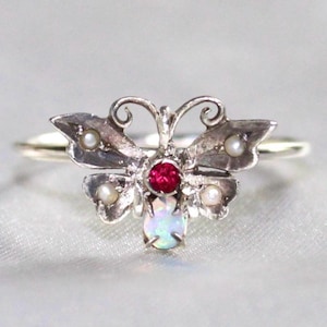 May include: A silver ring featuring a butterfly-shaped centerpiece. The butterfly is adorned with a central round red gemstone, a white oval opal at the base, and four small white pearls set within the wings. The metal has a polished finish.