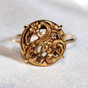 Puede incluir: Un anillo de tono dorado con una pieza central circular de diseño calado intrincado. La cara redonda muestra motivos florales en relieve y patrones de volutas, asegurada por cuatro pequeñas garras en una banda fina y pulida.