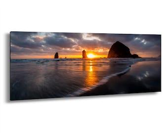May include: Panoramic canvas print of a sunset over a beach. The sky is filled with dark, dramatic clouds, with the sun breaking through in a burst of orange and yellow. The ocean reflects the colors of the sky, and several rock formations are visible.