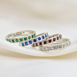 May include: Four eternity bands with different coloured gemstones. The bands are stacked on top of each other. The top band is set with clear diamonds, the next band is set with red rubies, the next band is set with blue sapphires, and the bottom band is set with green emeralds.