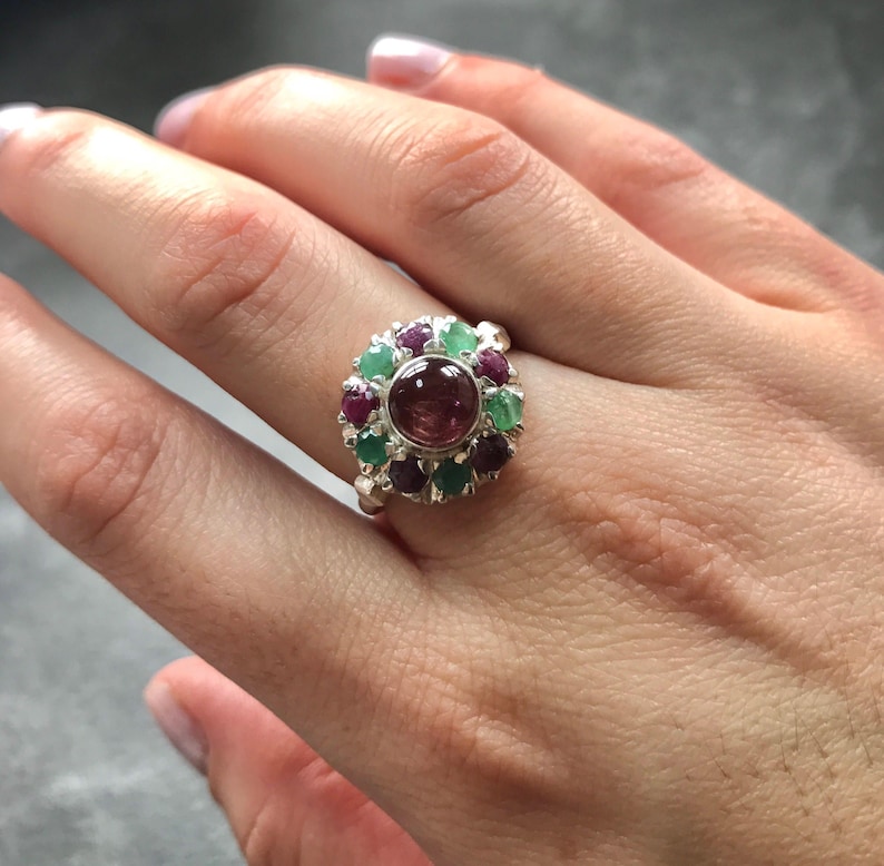 May include: A silver ring featuring a large, round, reddish-brown gemstone at the center, surrounded by smaller, alternating red and green gemstones. The ring is set on a hand.