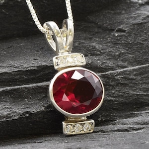 May include: A silver pendant necklace with a large, round, red gemstone surrounded by smaller, clear gemstones. The pendant is on a silver chain.