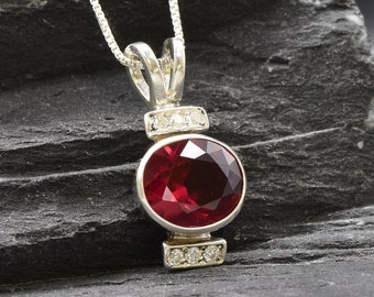 May include: A silver pendant necklace featuring a large, round, red gemstone surrounded by smaller, clear gemstones. The pendant hangs from a silver chain.