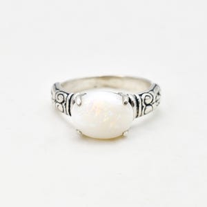 May include: A silver ring with an oval, iridescent white opal gemstone. The band has an ornate, swirling design on the sides. The opal is held in place by four prongs. This ring is a piece of jewellery.