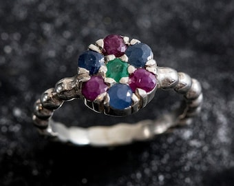 Op de afbeelding: Een zilveren ring met een cluster van edelstenen. De ring heeft een centrale opstelling van edelstenen, waaronder rode robijnen, blauwe saffieren en een groene smaragd. De band heeft een getextureerd, kralenontwerp. De ring is geschikt voor dagelijks gebruik.