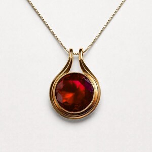 Könnte beinhalten: Eine Goldhalskette mit einem runden, facettierten, tiefroten Edelstein in einer polierten Goldfassung. Der Anhänger hängt an einer zarten Goldkette und zeigt ein klassisches Design mit einer glatten, geschwungenen Öse, die beide Teile verbindet.
