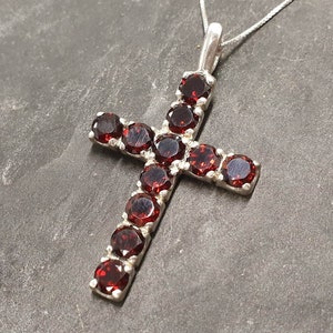 Puede incluir: Un colgante de cruz de plata con piedras preciosas rojas. La cruz está adornada con piedras redondas rojas engastadas en plata.