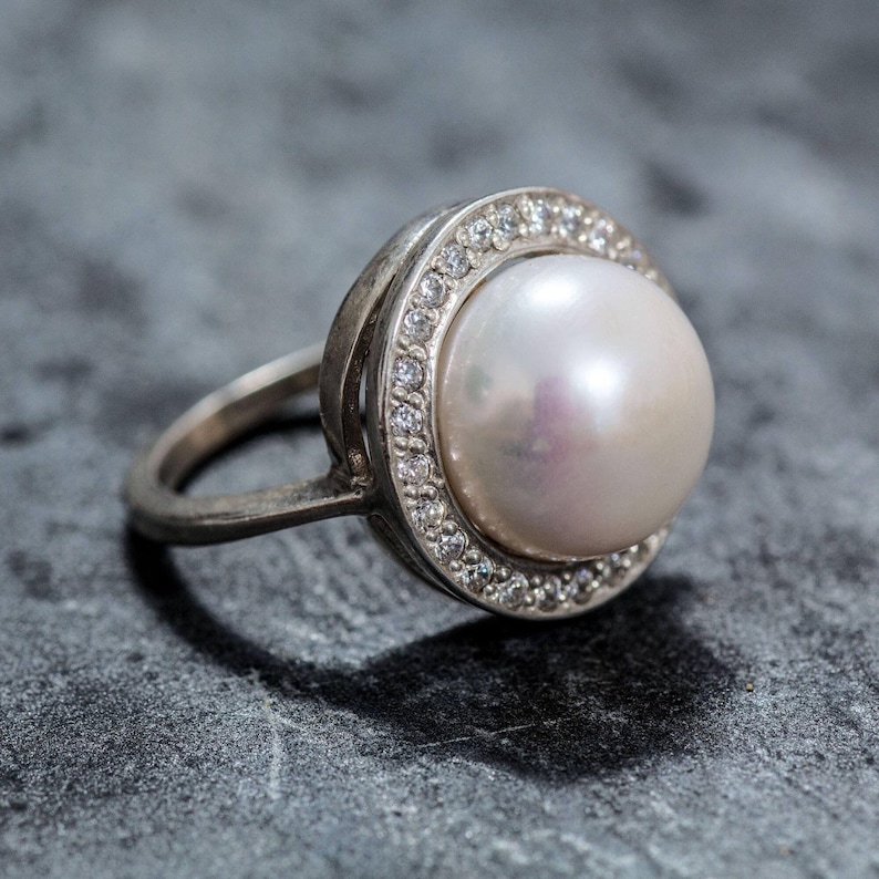 May include: A silver ring with a large white pearl set in a round bezel with small clear stones around the edge.