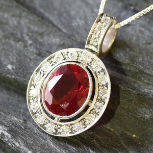 Könnte beinhalten: Eine silberne Halskette mit einem Anhänger, der einen großen ovalen roten Edelstein mit einem Heiligenschein aus kleineren weißen Edelsteinen zeigt.