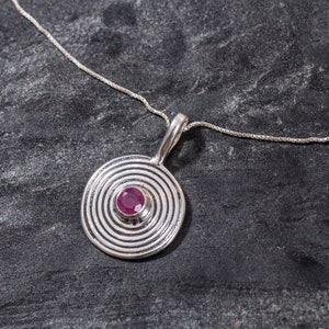 Peut inclure: Un collier en argent avec un pendentif circulaire orné d'un motif de cercles concentriques et d'une petite pierre précieuse rose au centre.