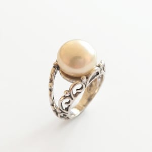 Puede incluir: Un anillo de plata con una perla grande, redonda y de color crema, montada sobre una banda con intrincados patrones de volutas caladas. El metal tiene un acabado pulido con &aacute;reas hundidas oscurecidas que resaltan el dise&ntilde;o de filigrana.