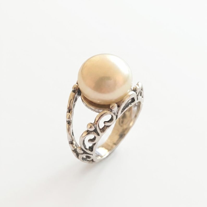 Puede incluir: Un anillo de plata con una perla grande, redonda y de color crema, montada sobre una banda con intrincados patrones de volutas caladas. El metal tiene un acabado pulido con &aacute;reas hundidas oscurecidas que resaltan el dise&ntilde;o de filigrana.