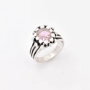 Peut inclure: Une bague en argent avec une pierre ovale rose sertie dans une bélière en argent texturée. La bague a une large bande avec trois lignes en relief.